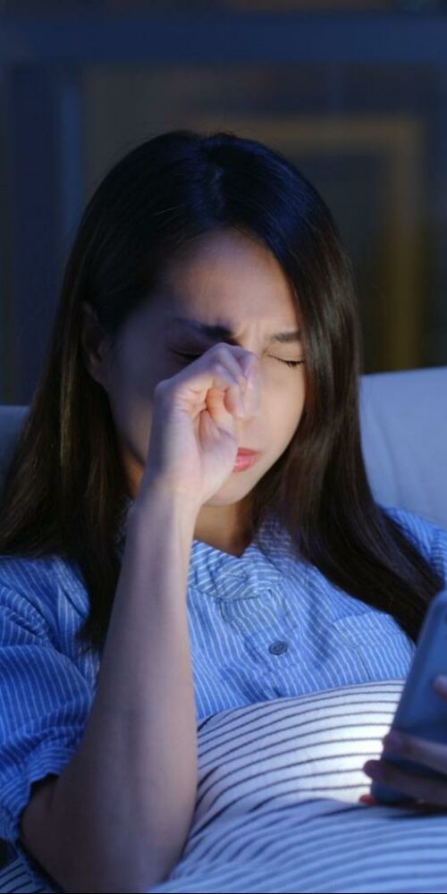 A woman sitting on a couch at night, rubbing her eyes while looking at her smartphone, indicating discomfort possibly due to dry eyes.