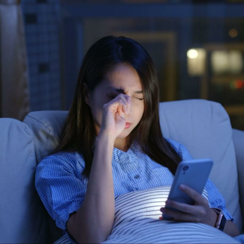 A woman sitting on a couch at night, rubbing her eyes while looking at her smartphone, indicating discomfort possibly due to dry eyes.