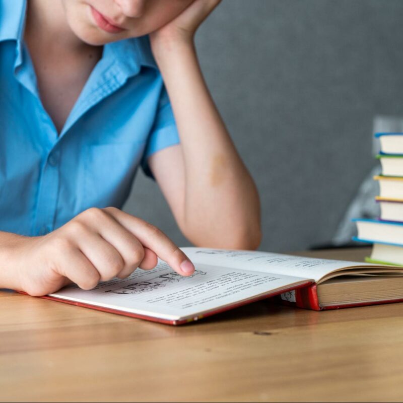 A student reading a book while pointing to the text, with a stack of books on the table, illustrating challenges in reading related to dyslexia.