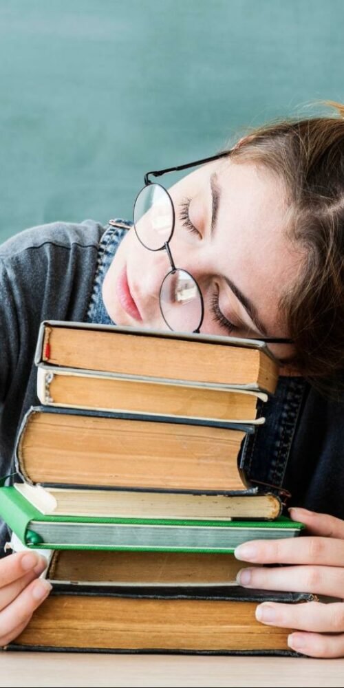 A student wearing glasses resting their head on a stack of books, looking tired and overwhelmed in a classroom.