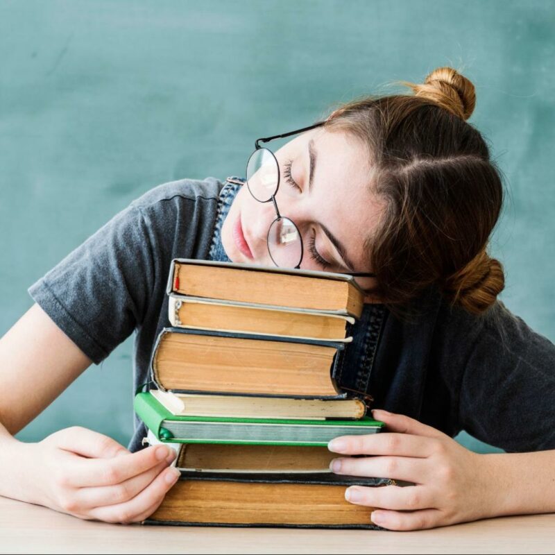 A student wearing glasses resting their head on a stack of books, looking tired and overwhelmed in a classroom.