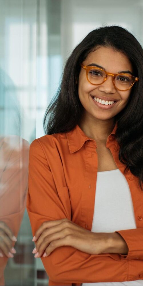 A young woman with long dark hair, wearing glasses and an orange shirt, smiling confidently while leaning against a glass wall in a bright office setting.