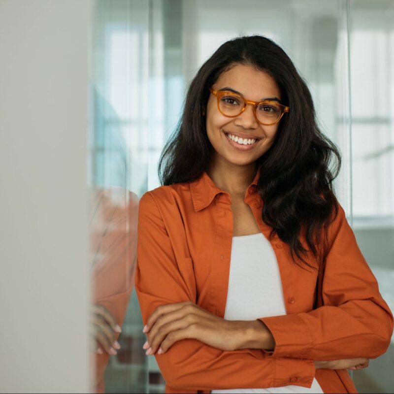 A young woman with long dark hair, wearing glasses and an orange shirt, smiling confidently while leaning against a glass wall in a bright office setting.